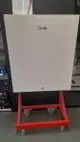 Danfoss-Gerät auf rotem fahrbarem Untergestell, mit einer glatten weißen Front. Im Hintergrund sind Werkzeuge und Kartons sichtbar.